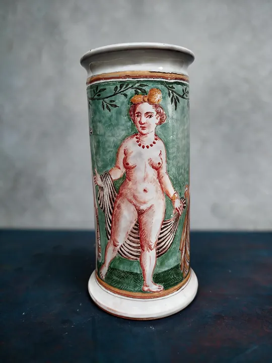 Auf dem hohen zylindrischen Gefäß steht eine schlanke Frauenfigur vor einem grünen Feld. Ein kleiner Vogel sitzt auf ihrem Kopf, daneben verlaufen rote Konturen und grüne Blätter über die helle Fläche.