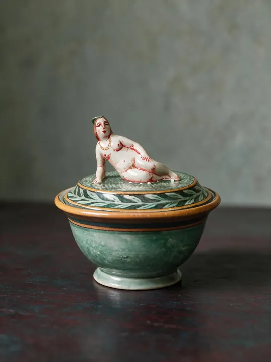 Vier Figuren ziehen sich rund um den bauchigen Gefäßkörper. Auf dem Deckel liegt eine Frauenfigur; grüne und rotbraune Bänder markieren Fuß und Schulter.