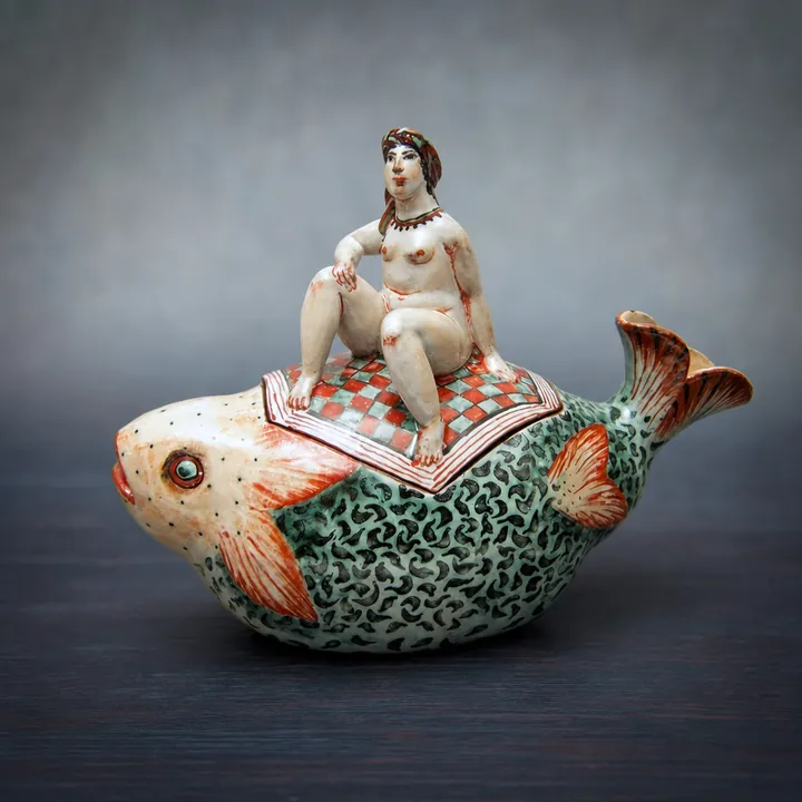 Die große rundliche Fischfigur trägt eine sitzende Frauenfigur auf einer rot-weiß karierten Decke. Grüne, schwarze, rote und helle Muster gliedern Schuppen, Flossen und Körper.