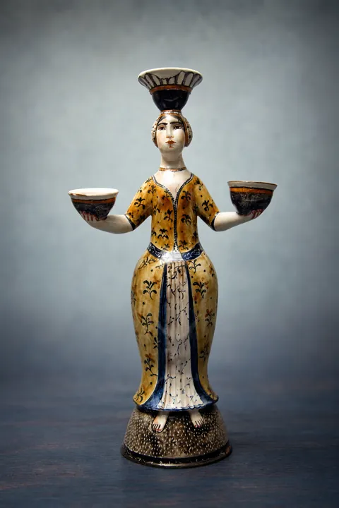 Die stehende Frauenfigur balanciert eine Schale auf dem Kopf und hält zwei weitere seitlich in den Händen. Das lange Gewand ist gelb, blau, weiß und schwarz gegliedert und steht auf einem dunklen Rundsockel.