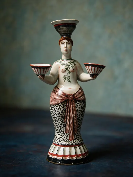 Die stehende Frauenfigur balanciert eine Schale auf dem Kopf und hält zwei weitere seitlich in den Händen. Weiß, Rotbraun, Schwarz und Grün bestimmen das symmetrische Dekor von Figur, Gewand und Sockel.