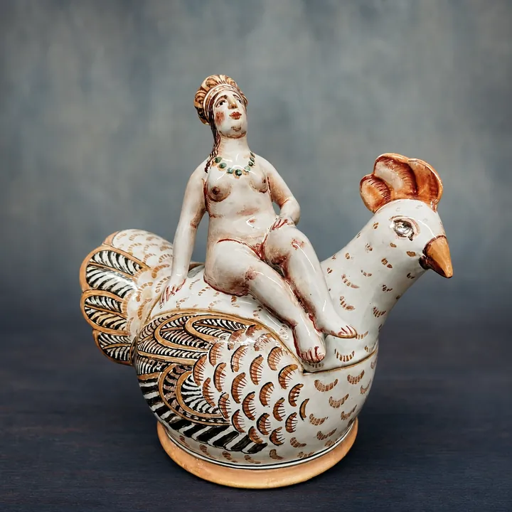 Eine sitzende Frauenfigur befindet sich auf dem Rücken eines weißen Hahns. Das Gefieder ist mit braunen, schwarzen und goldfarbenen Linien und Punkten dicht verziert.
