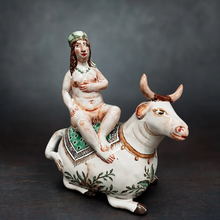 Eine sitzende Frauenfigur befindet sich auf dem Rücken eines liegenden weißen Stiers mit dunklen Hörnern. Grüne Pflanzenmotive sowie rote, schwarze und goldbraune Akzente schmücken die Figurengruppe.