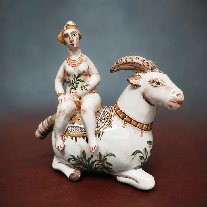 Eine sitzende Frauenfigur befindet sich auf einem liegenden weißen Widder mit eingerollten Hörnern. Grüne Pflanzenmotive und braune, rote sowie schwarze Details akzentuieren die helle Oberfläche.