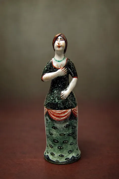 Die schlanke stehende Frauenfigur trägt ein dunkles Oberteil und einen langen grünen Rock. Rote Bänder, schwarze Punktmuster und eine grüne Halskette setzen farbige Akzente.