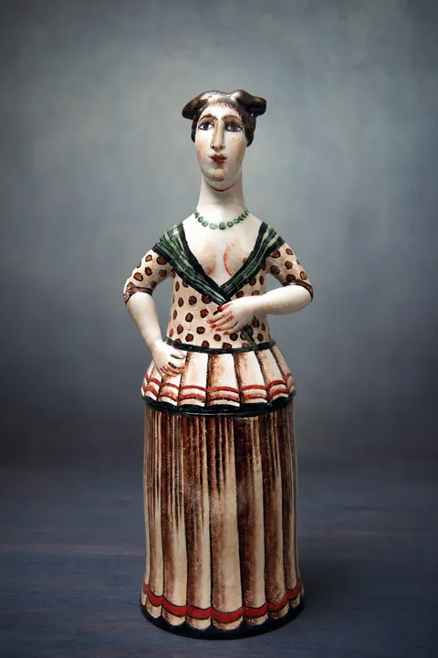 Die stehende Frauenfigur trägt einen langen senkrecht gestreiften Rock und ein weißes Oberteil mit dunklen und roten Bändern. Ihre Hände liegen symmetrisch vor der Taille.