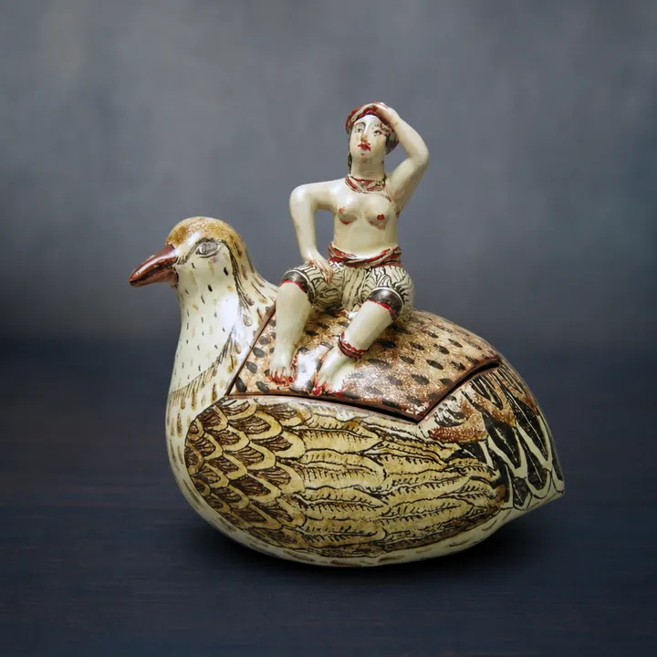 Die rundliche Vogelfigur trägt eine sitzende Frauenfigur auf dem Rücken. Gelbbraune, schwarze, grüne und rote Linien bilden ein dichtes Feder- und Flächendekor.
