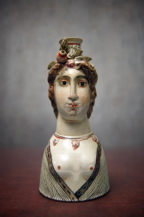 Die weibliche Büste zeigt ein helles Gesicht, dunkles lockiges Haar und einen erhöhten floralen Kopfschmuck. Das Gewand ist in Grau, Schwarz und Weiß mit feinen Linien gegliedert.