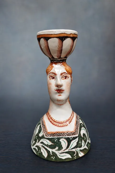 Die weibliche Büste trägt eine kelchförmige Schale auf dem Kopf. Gesicht, Kleidung und Schale sind in Weiß, Dunkelgrün, Rotbraun und Schwarz ornamental gestaltet.