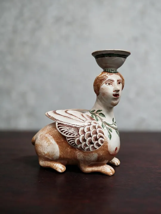 Ein gedrungener Hasenkörper mit langen Ohren trägt einen Frauenkopf und eine kleine Schale. Auf der hellen Fläche stehen braunrote Linien, Punkte und ein netzartig gemusterter Kragen.