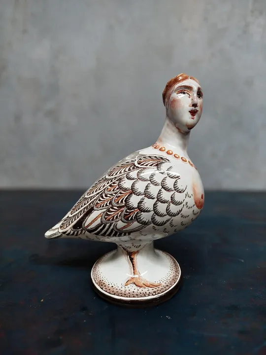 Ein Vogel mit aufgerichtetem Frauenkopf steht auf einem runden Fuß. Schuppen, Bögen und Linien formen ein braun-schwarzes Gefieder, rotbraune Akzente liegen an Gesicht und Hals.