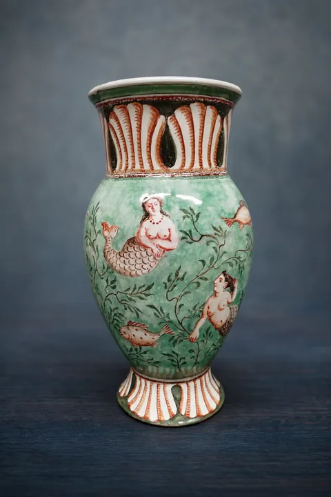 Die bauchige grüne Vase steht auf einem schmalen Fuß und besitzt einen weit geöffneten Hals. Rötliche Figurenmotive, dunkle Blattranken und senkrechte Streifen gliedern die Oberfläche.