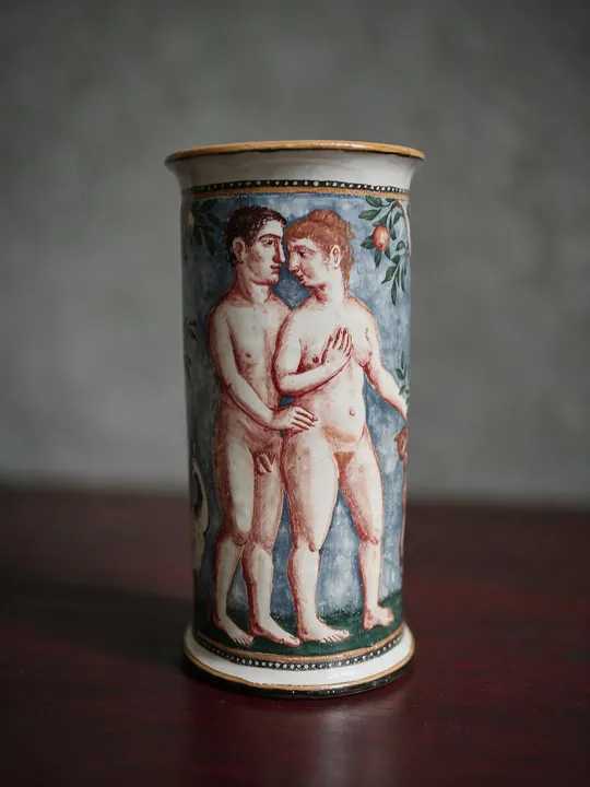 Ein unbekleidetes Figurenpaar steht dicht beieinander auf der hohen zylindrischen Vase. Rotbraune Konturen liegen vor grünem Grund, waagerechte Bänder schließen das Bildfeld ein.