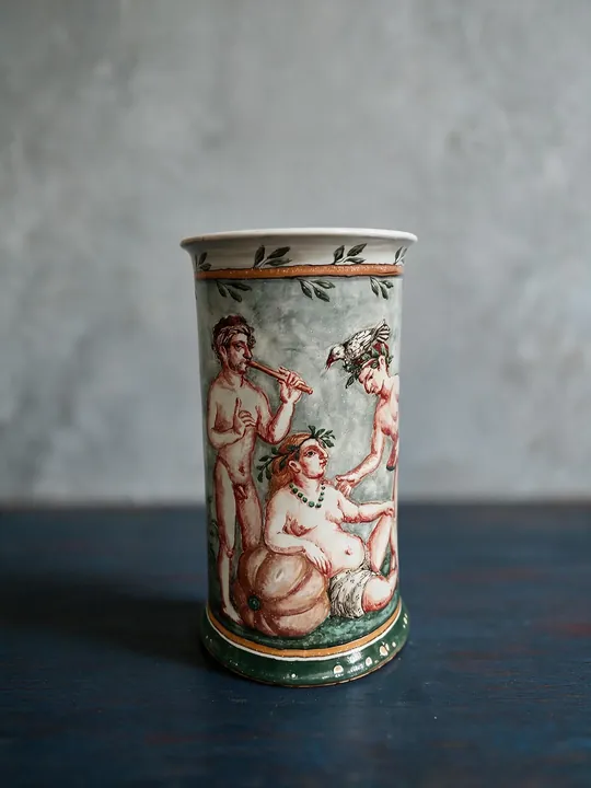 Auf der hohen zylindrischen Vase steht ein Paar zwischen Zweigen und Blättern. Rotbraune Konturen, grüne Flächen und schmale Bänder begrenzen das Bildfeld.