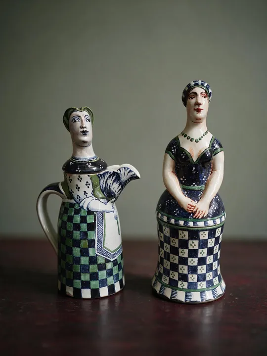 Zwei stehende Frauenfiguren bilden eine Gruppe, eine von ihnen hält einen kleinen Vogel. Schwarz-weiße Schachbrettfelder, dunkelblaue Partien und grüne Akzente laufen über Gewänder und Sockel.