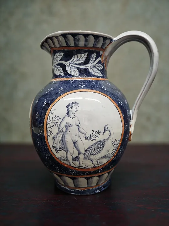In einem hellen Medaillon steht eine Frauenfigur mit ausgebreiteten Vogelflügeln. Kobaltblaue Ornamente, braune Konturen und ein großer Rundhenkel geben der Vase ihre Form.