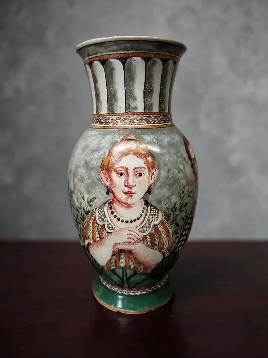 Ein Frauenporträt mit rotem Haar nimmt die Vorderseite der bauchigen Vase ein. Grüne Blätter und Bänder, schwarze Konturen und ein durchbrochener Hals umgeben das Gesicht.