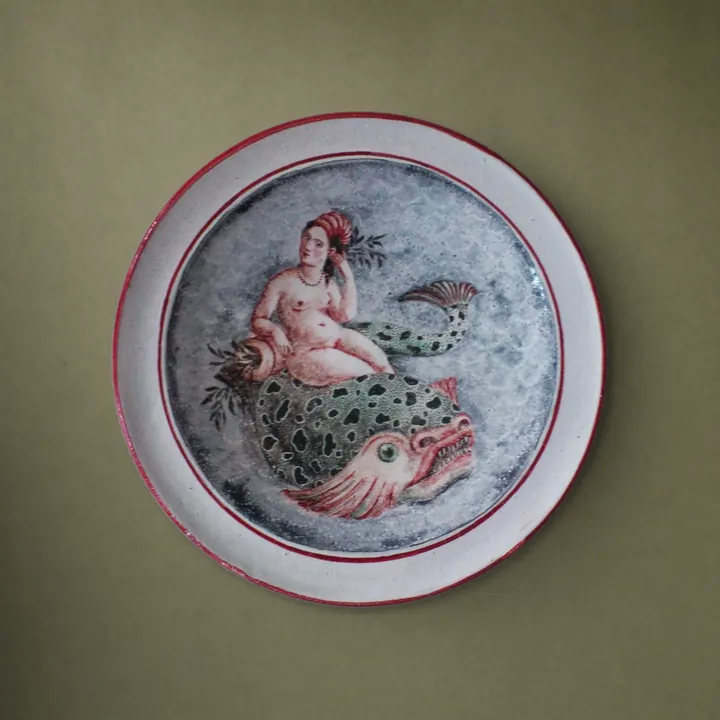 Eine unbekleidete Frauenfigur sitzt auf einem großen gefleckten Fisch. Das helle Rundbild wird von zwei feinen rotbraunen Linien umrandet.