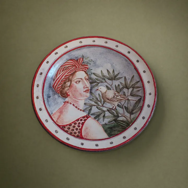 Ein Frauenkopf mit roter Kopfbedeckung wendet sich einem kleinen Vogel zwischen belaubten Zweigen zu. Rote Punkte und Linien ziehen einen Rand um das Bild.