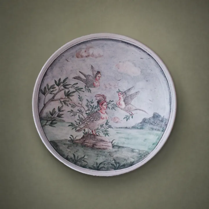 Mehrere geflügelte Figuren bewegen sich zwischen Wasser, Bäumen und Gräsern. Grau, Grün und Rosa bestimmen die helle Landschaft auf dem Teller.